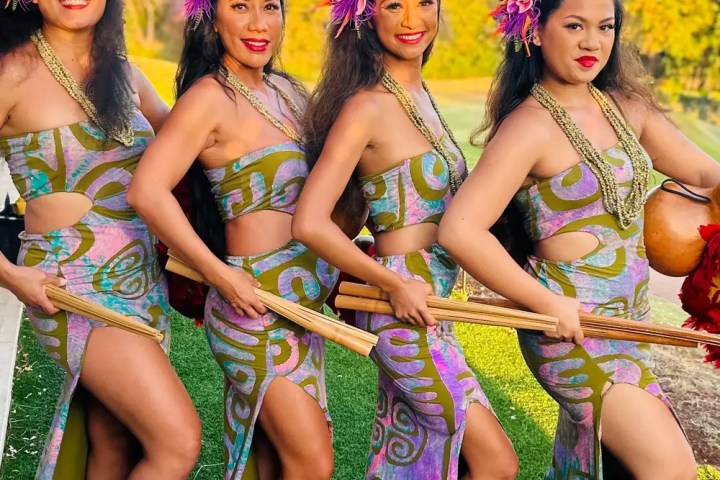 Four women in colorful floral outfits and headpieces, posing with a traditional dance pose outdoors.