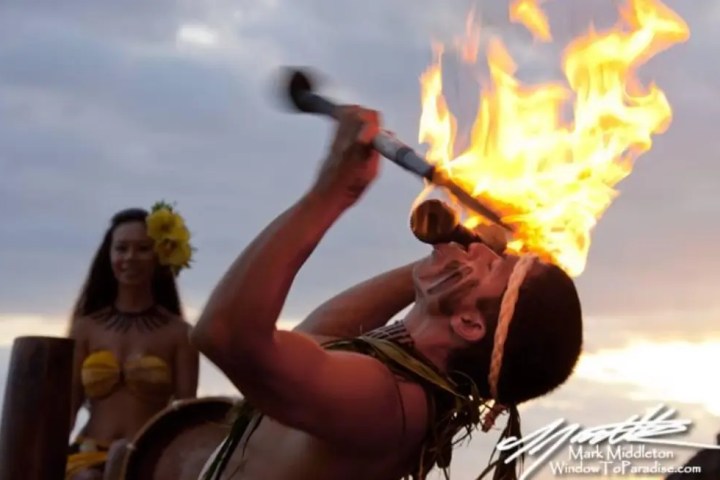 Performer blows fire from a conch shell, with a woman in traditional attire in the background.