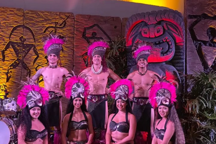 Group in colorful traditional costumes posing on stage with tropical backdrop.