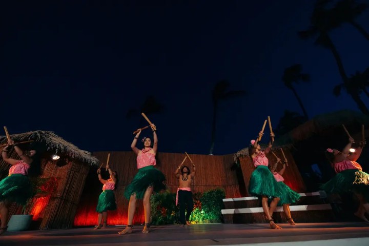 Dancers in skirts perform with sticks on an outdoor stage at night.