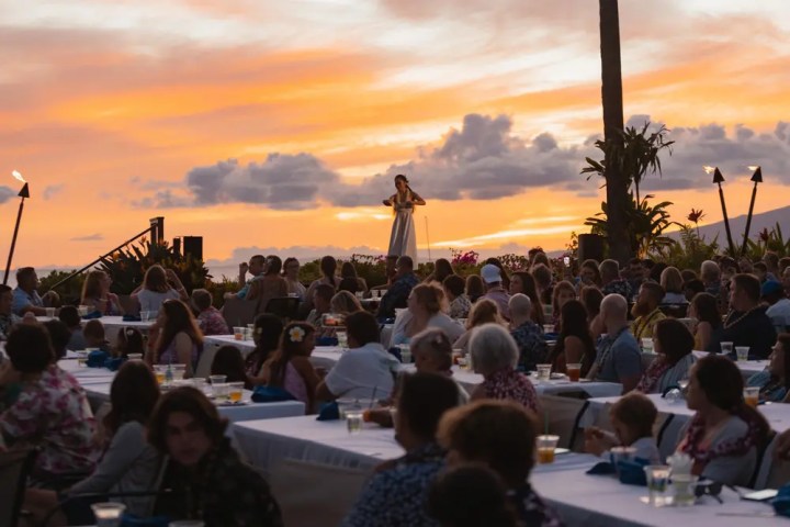 Outdoor event at sunset with a performer onstage and audience seated at tables.