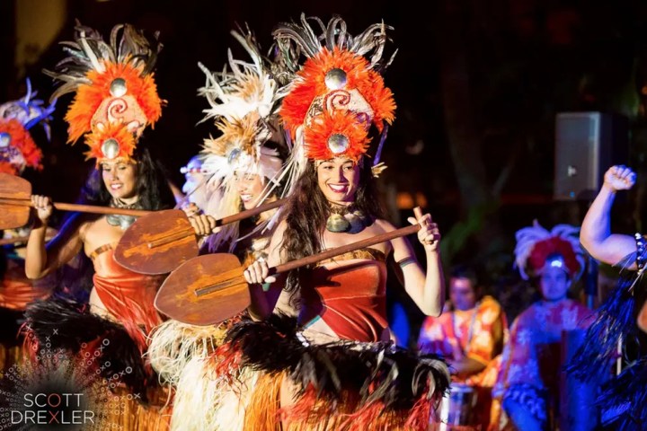 Dancers in vibrant costumes and feathered headdresses perform with paddles.