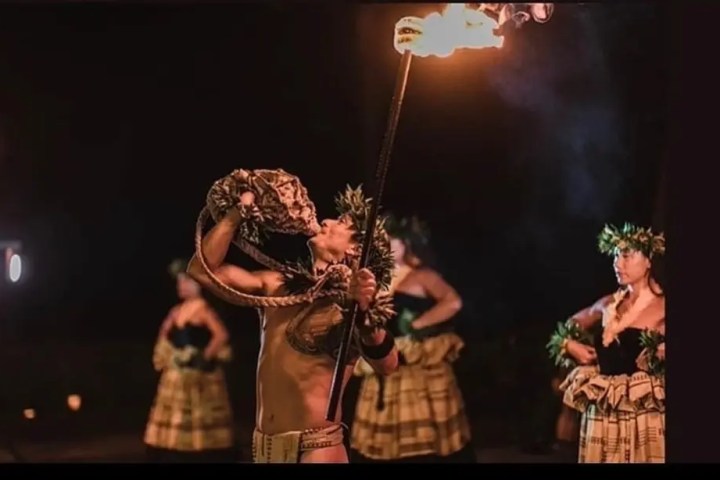 Performer blowing conch shell, holding flame, with dancers in background.