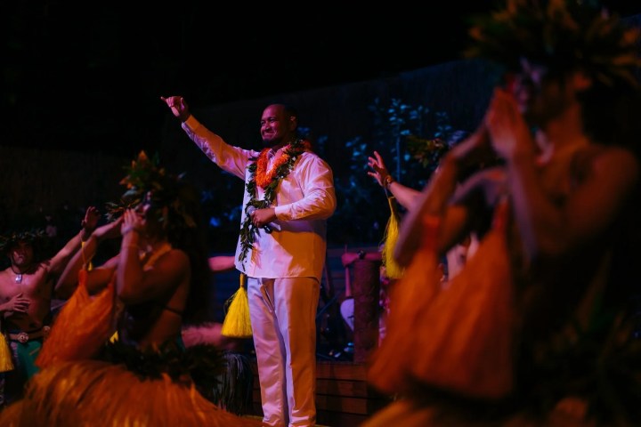 Person in white suit with leis stands among hula dancers at night.