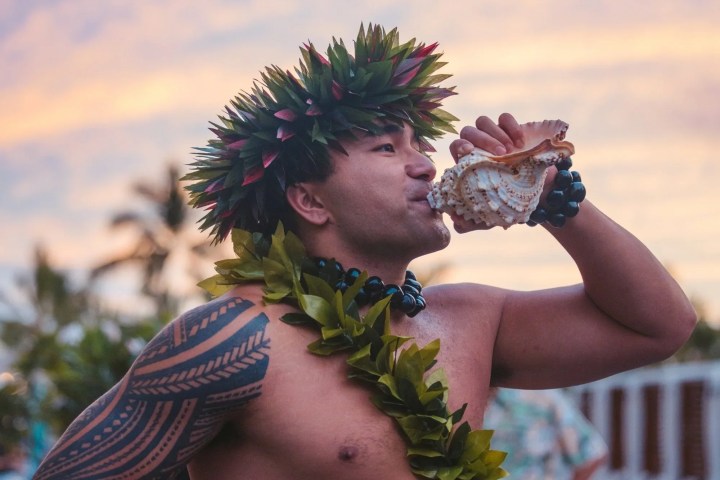 Man in traditional attire blowing a conch shell at sunset.