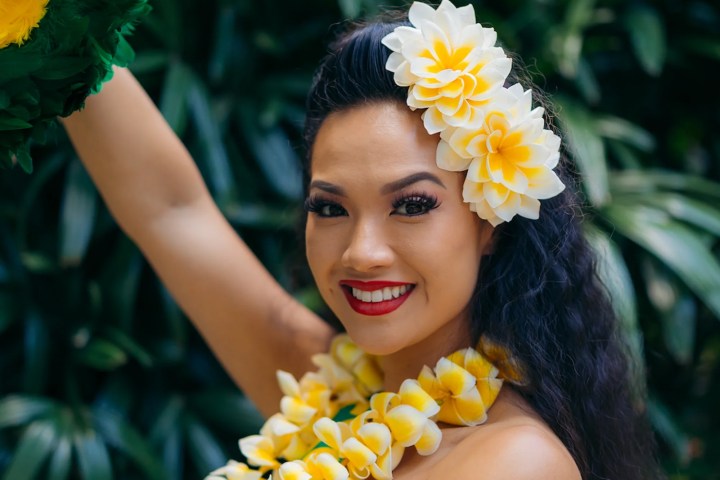 Woman in floral dress with lei and flower crown, smiling outdoors.