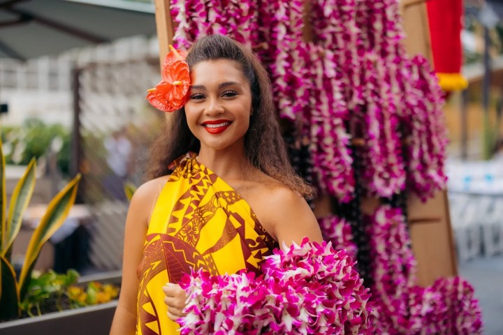 Smiling woman in tropical dress and lei with flower in hair, background of purple leis.